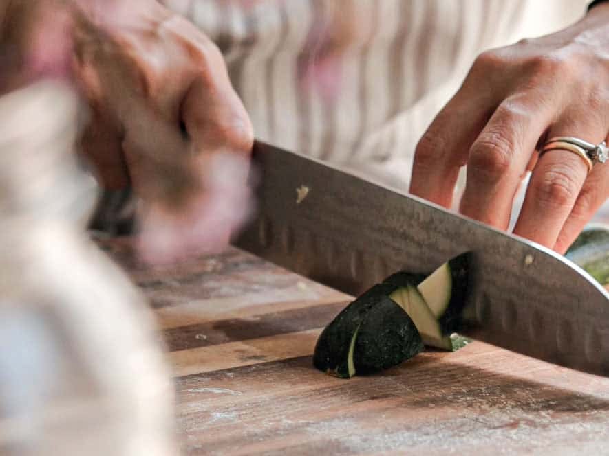 Close-up of hands slicing a zucchini on a wooden cutting board. One hand holds the zucchini steady while the other uses a knife to cut. The person is wearing a ring on their left hand.